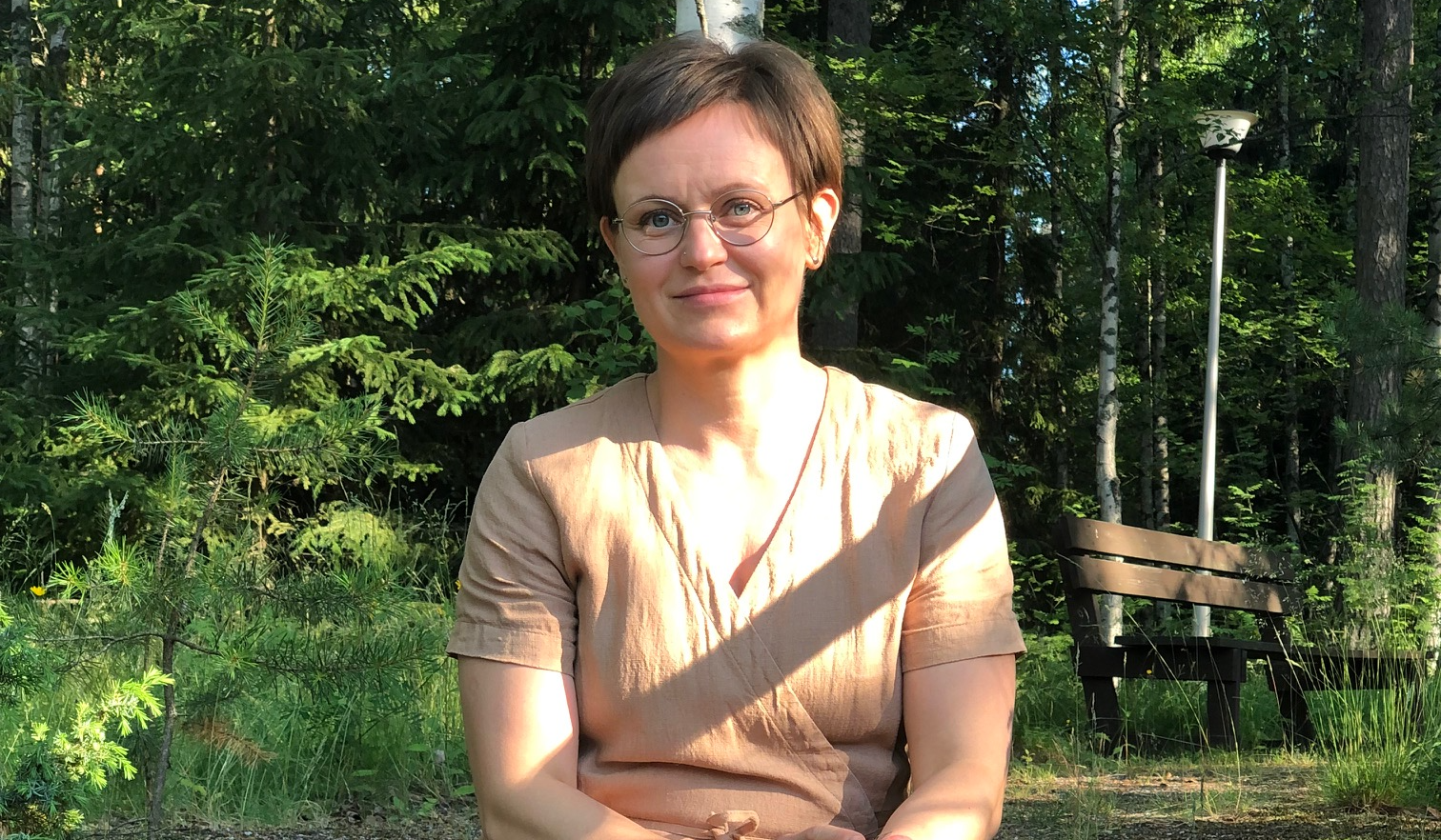 Sanna Suuripää hymyilee. Taustalla vihreä puisto, penkki ja valopylväs.